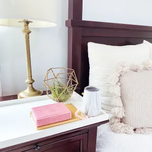 A bedside table with a decorative geometric planter, a pink sparkly book, a mug, and a brass lamp.