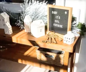 A wooden shelf decorated with miniature white houses, a felt letter board saying 'Baby It's Cold Outside', vintage keys, and evergreen sprigs.