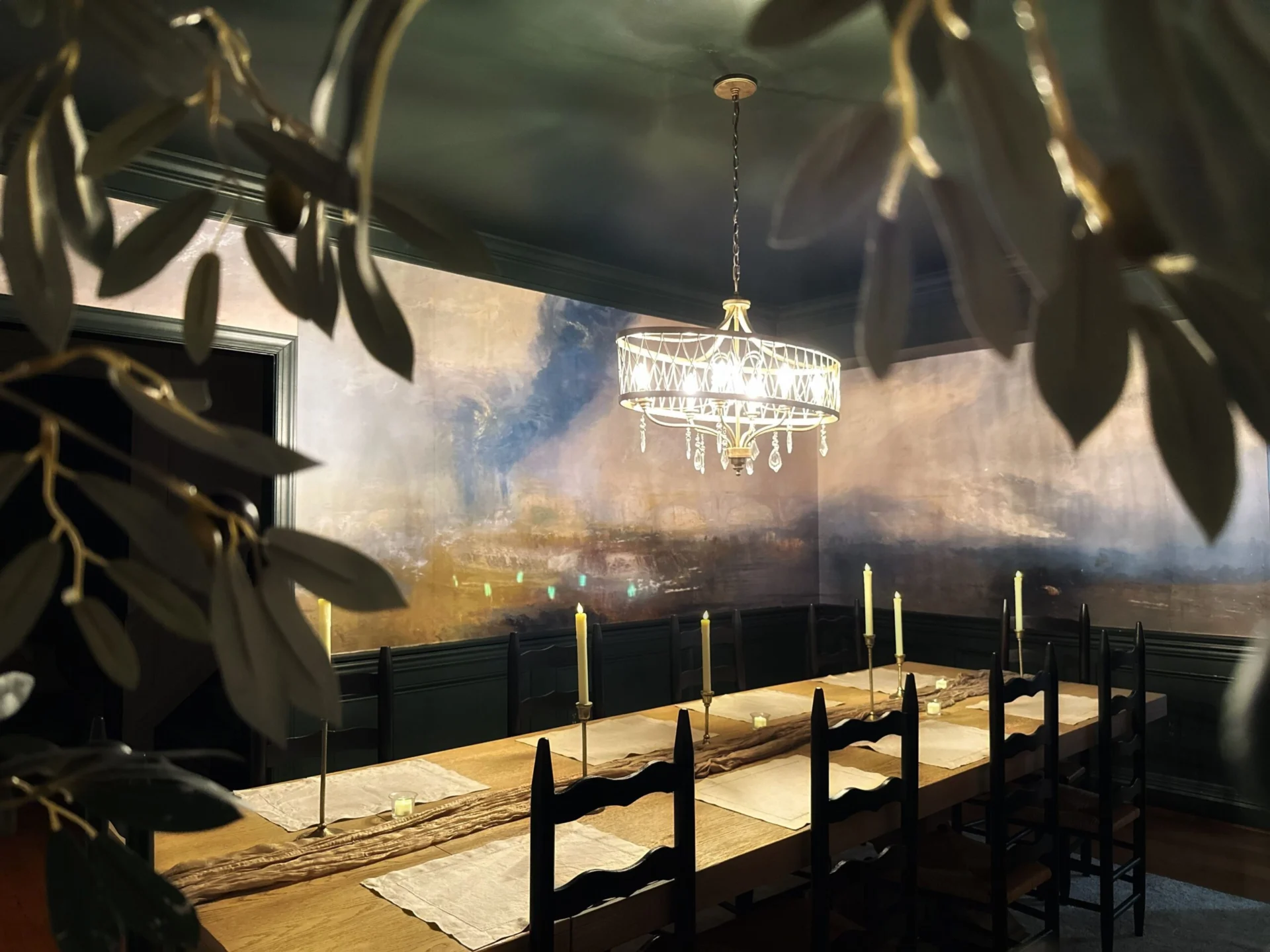 A long wooden dining table set for a meal with candles and a textured runner, illuminated by a crystal chandelier, in front of a dramatic cloudy mural.
