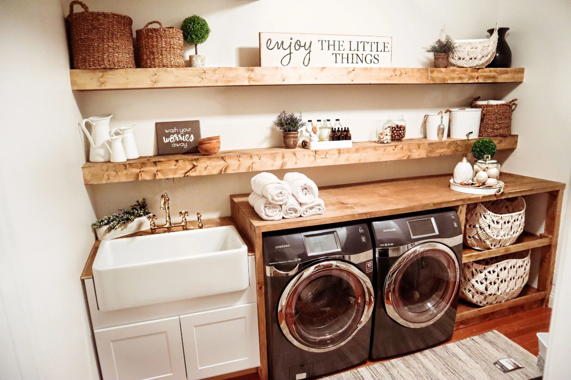 A clean and organized farmhouse laundry room featuring a white apron sink, two dark Samsung washers, and wooden shelving filled with baskets, decor, and linens.