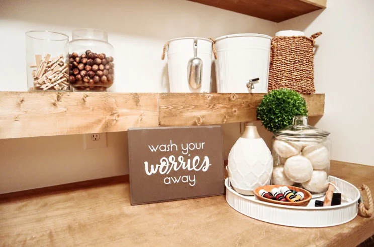 Rustic wooden shelves in a laundry room with decorative jars, white buckets, and a sign that says 'wash your worries away'.
