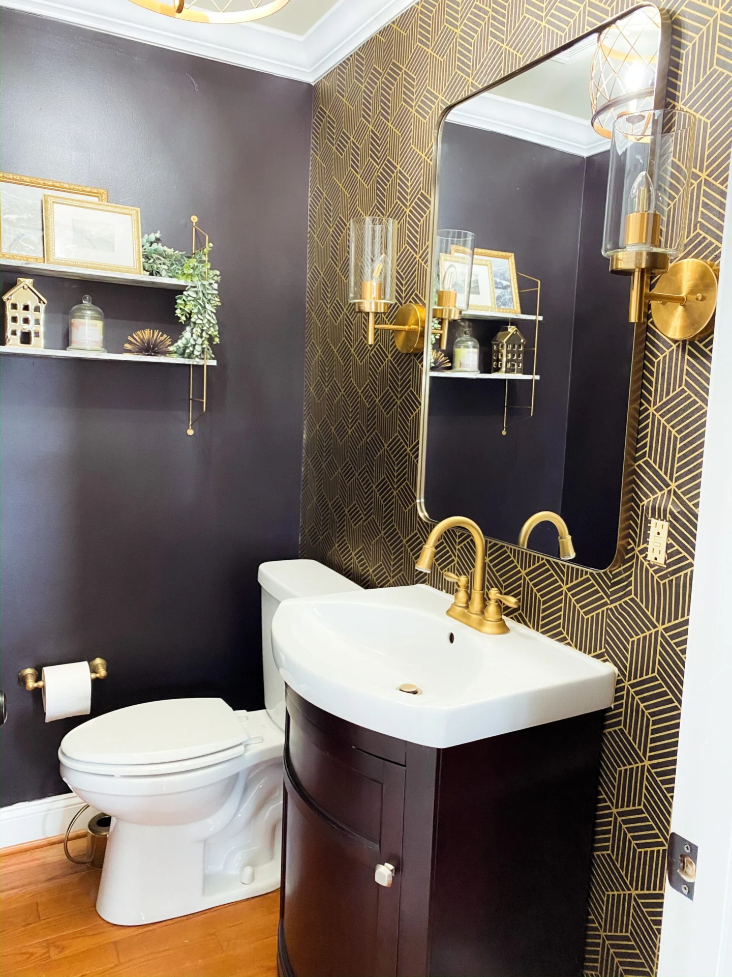 A small powder room featuring dark walls, gold geometric wallpaper, a white vanity with a gold faucet, and a white toilet.