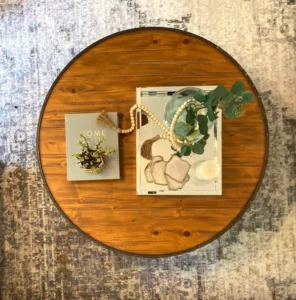 Overhead view of a round wooden coffee table with decorative items including a book, vase with greenery, beads, and coasters.