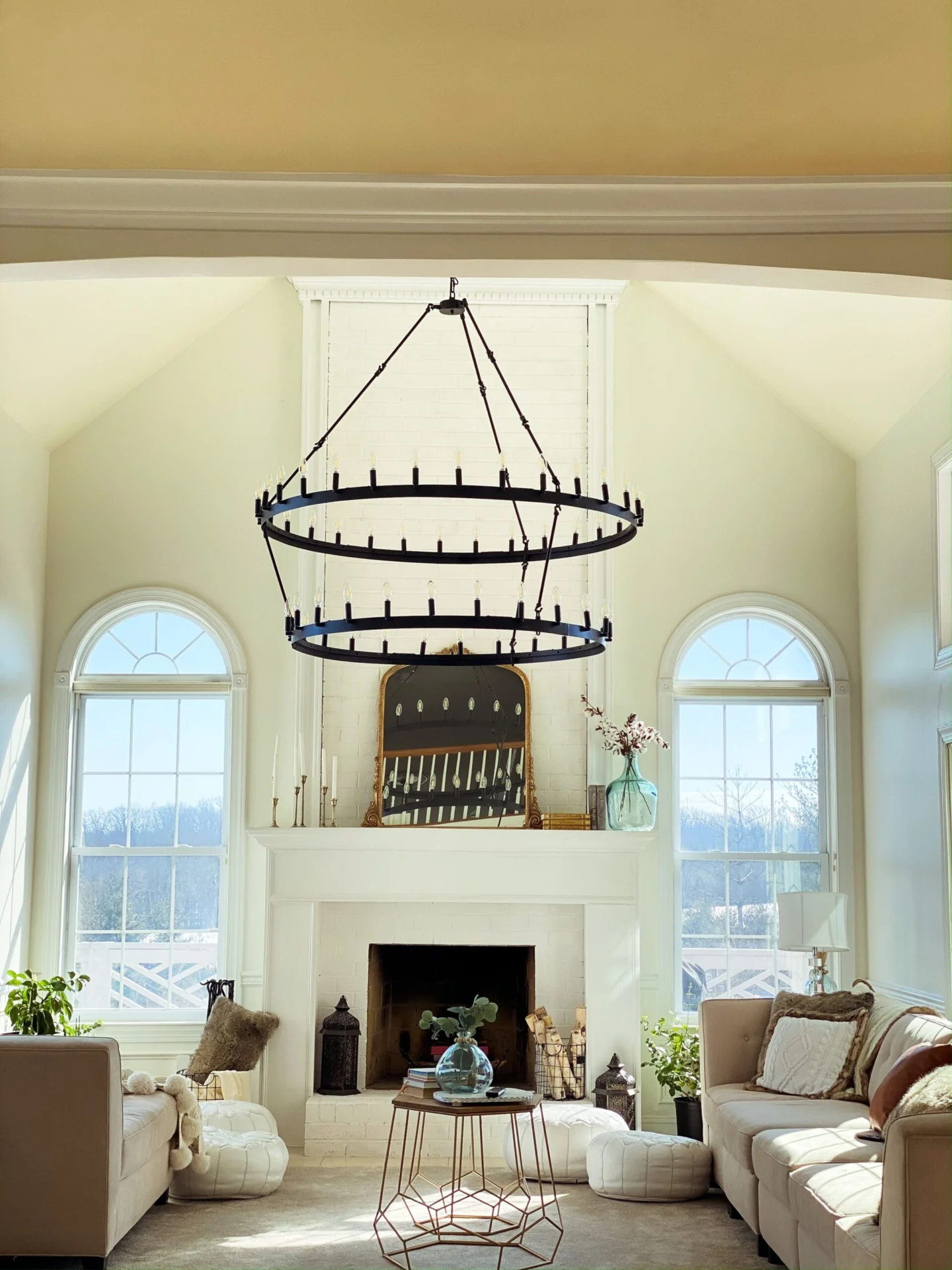 A bright and airy living room featuring a grand fireplace, a large two-tiered black chandelier, and two arched windows.