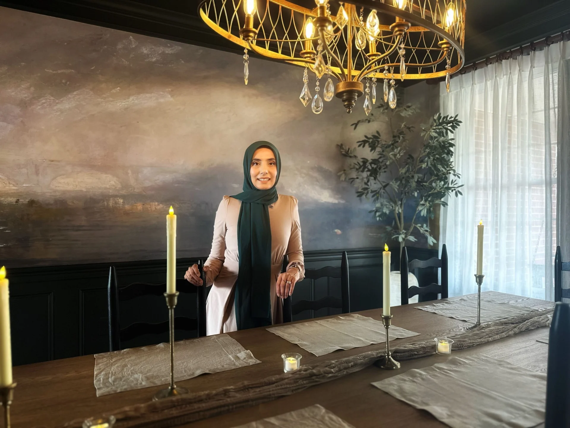 A woman wearing a green hijab and a light pink dress stands at a wooden dining table set with placemats, runners, and candles. She is smiling and leaning on a chair, with a large dramatic landscape painting on the wall behind her.