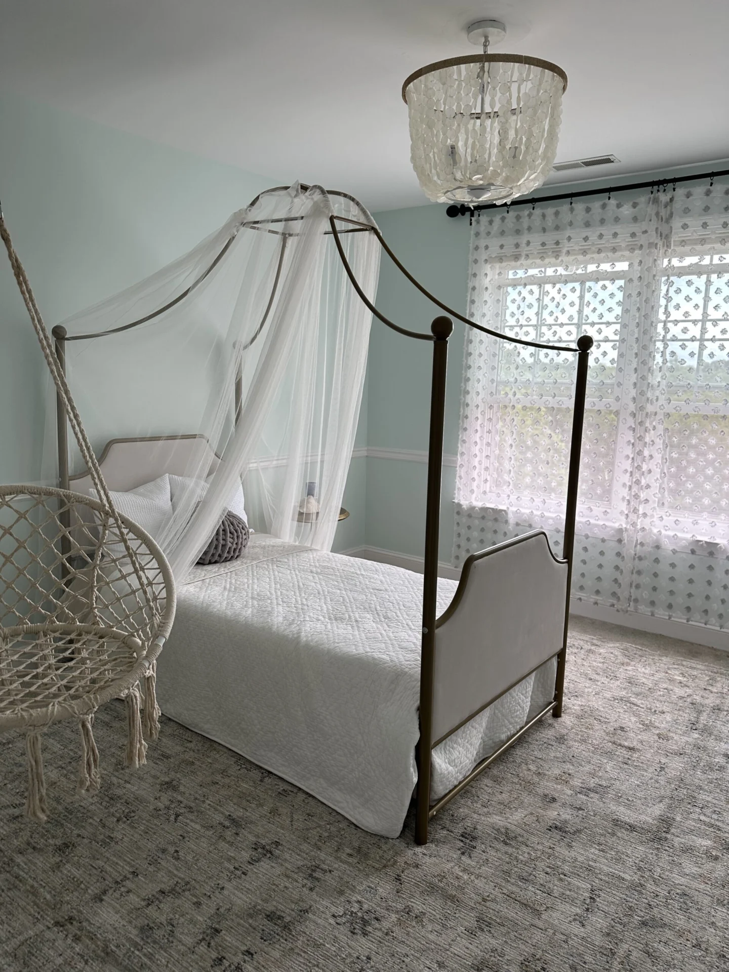 A bright and airy bedroom featuring a four-poster canopy bed with sheer white drapes, a macrame hanging chair, a patterned rug, and light blue walls.