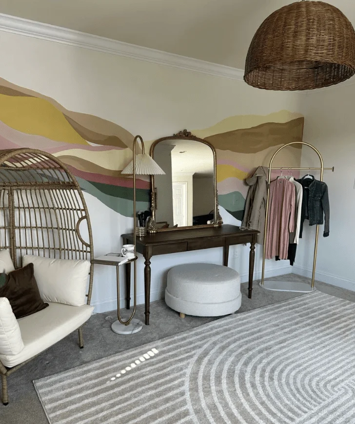 A bohemian bedroom with a unique abstract mural on the wall, featuring a cane egg chair, a dark wood vanity with a mirror, a plush ottoman, and a gold clothing rack.
