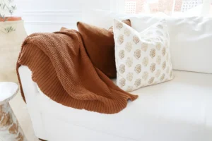 Close-up of a white sofa with a chunky knit rust-colored throw blanket draped over the arm, next to two decorative pillows: one solid brown velvet and another with a beige floral pattern on a white background.