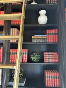 A tall, dark wooden bookshelf with a golden ladder. The shelves are filled with red and gold hardcover books, a white vase, and decorative items.